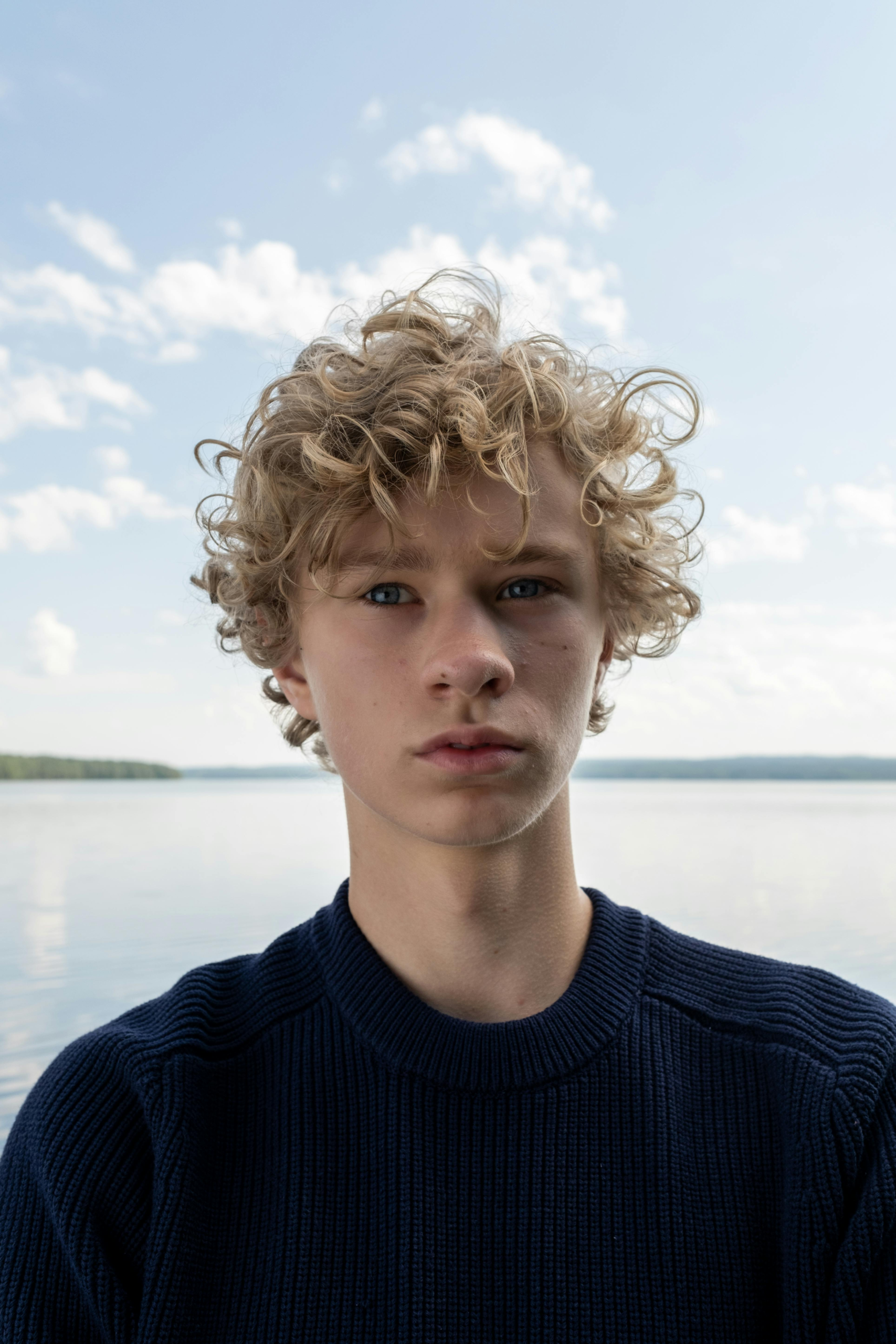 Close-up portrait of a blond teenager with curly hair at a lake, daytime.